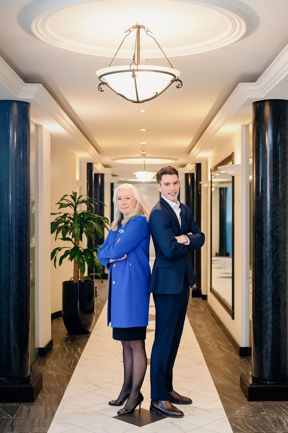 A woman and a man stand back-to-back in a luxurious hallway with arms crossed. Senior financial planner and principal, Patricia Howard in a blue blazer and Charles Osmond with short, dark hair and is wearing a dark navy suit with a white shirt.