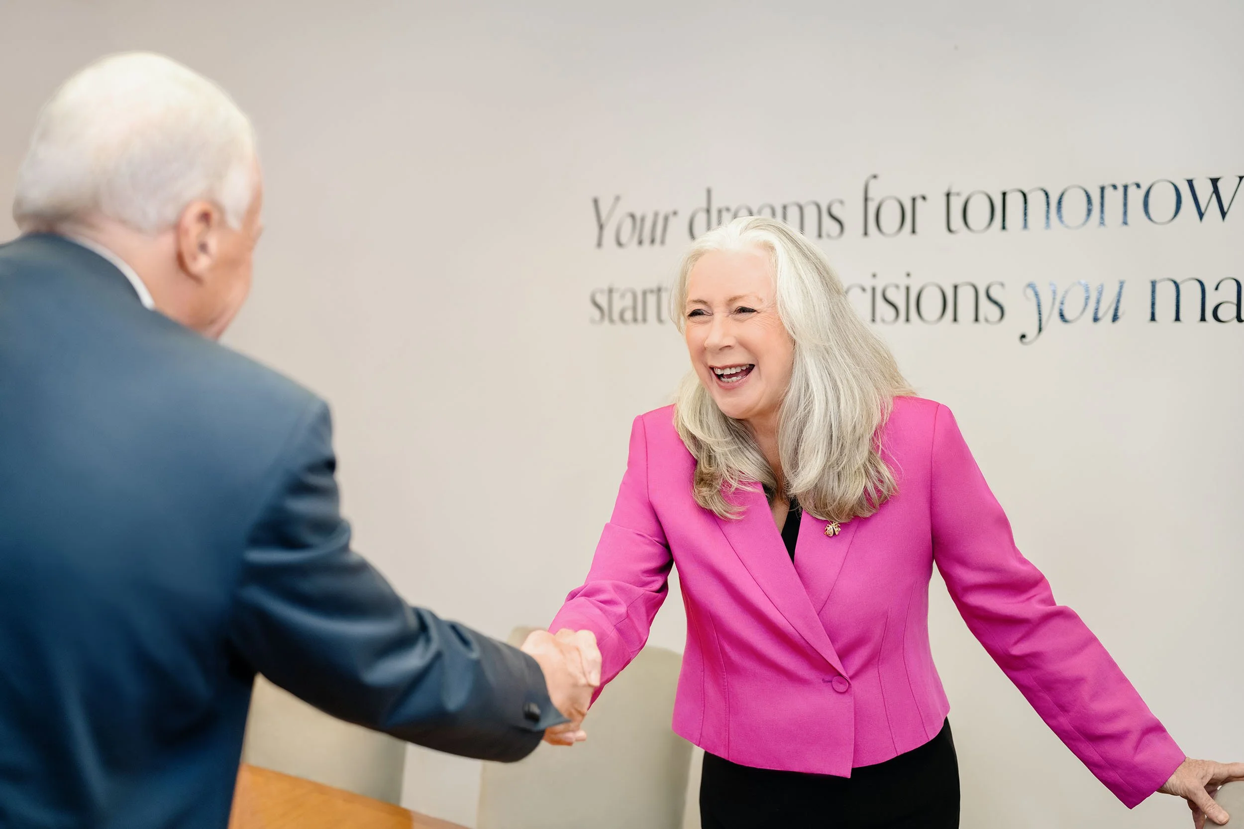 Senior financial planner and principal, Patricia Howard  with long gray hair wearing a pink blazer smiling and shaking hands with an older man in a suit, in their Kew, Melbourne office setting.