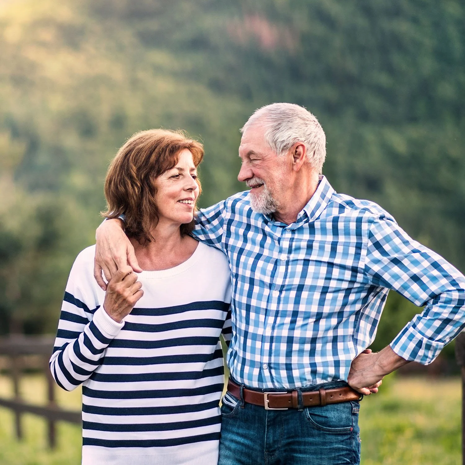 A middle-aged couple smiling at each other outdoors with a blurred green background.