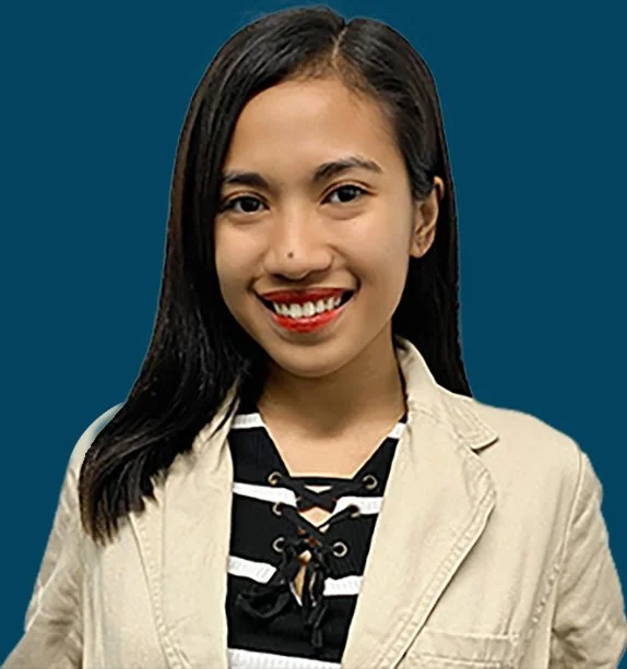 A young woman with long dark hair, smiling, dressed in a beige blazer over a black and white striped top with a lace-up neckline, against a solid blue background.