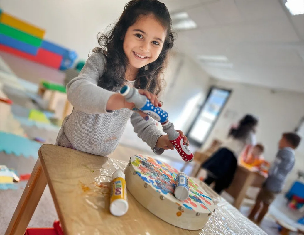A young girl with curly dark hair smiling at the camera while holding a toy hammer in a classroom. There are children and adults in the background, and colorful educational materials on the table.