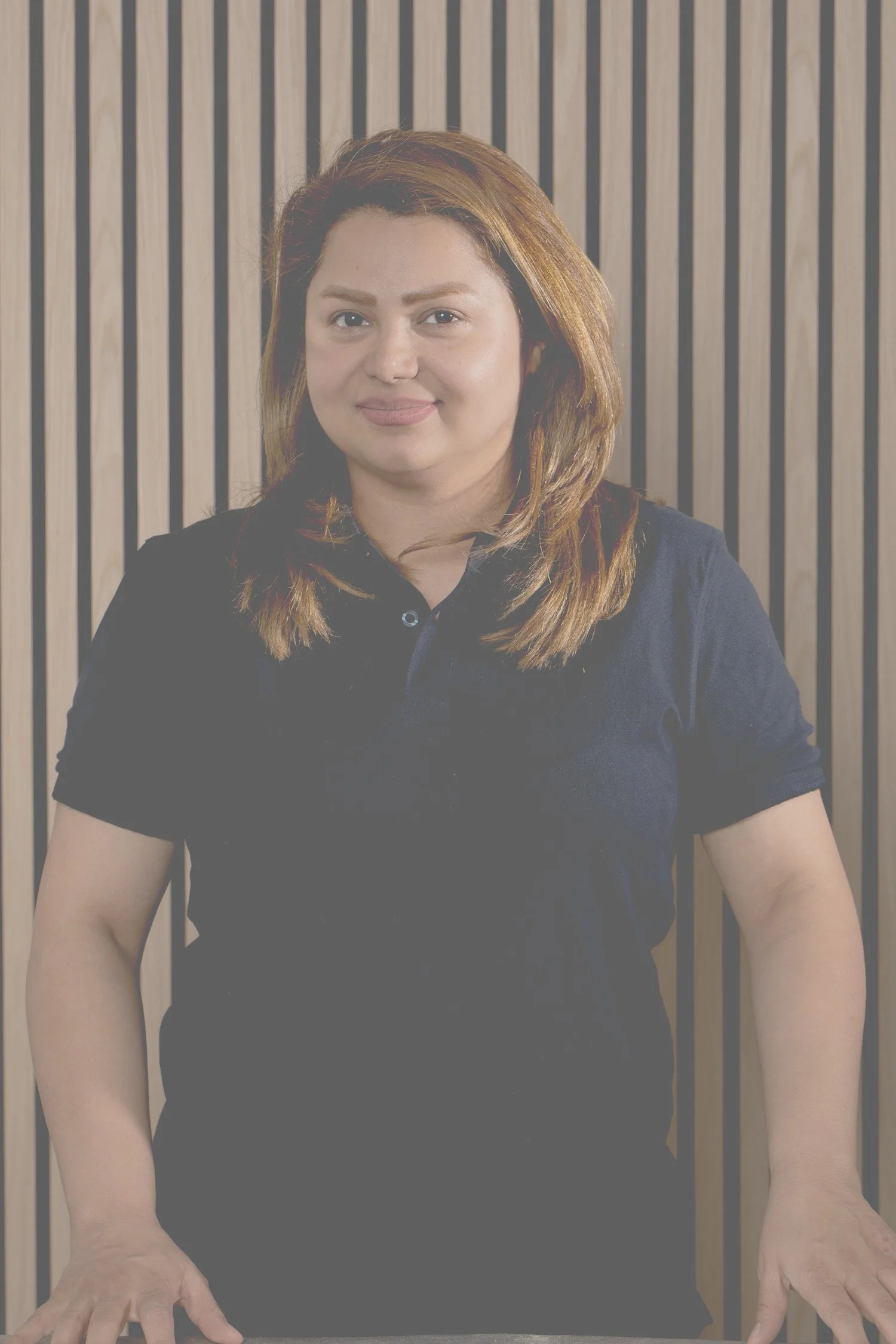 Eine Frau mit braunen, leicht gewellten Haaren steht vor einer Wand mit vertikalen Holzlamellen. Sie trägt ein dunkelblaues Polohemd und schaut in die Kamera.