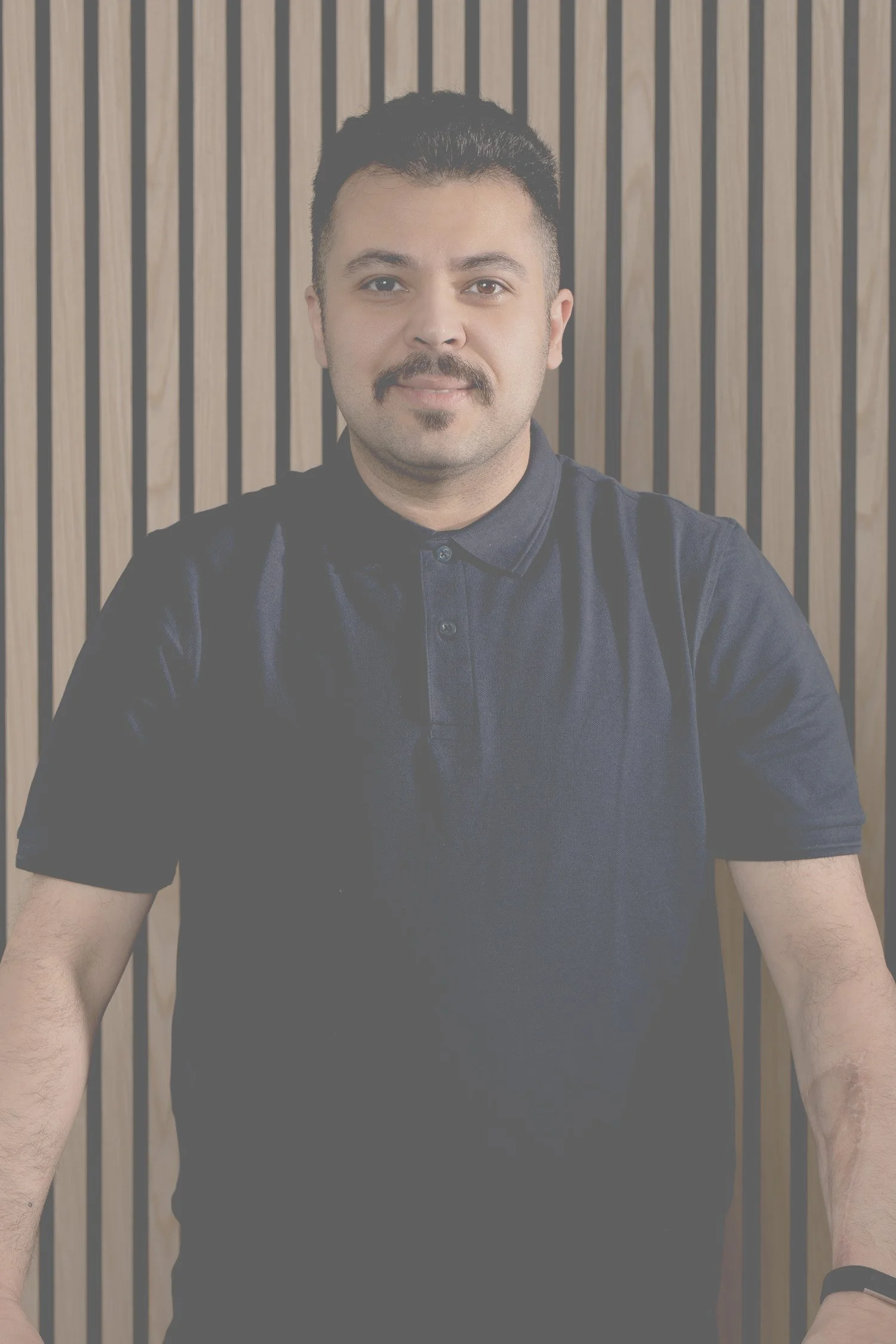 Porträt eines Mannes mit dunklem Haar, Schnurrbart und Bart, trägt ein dunkles Polo-Shirt, steht vor einer Holzvertäfelung.