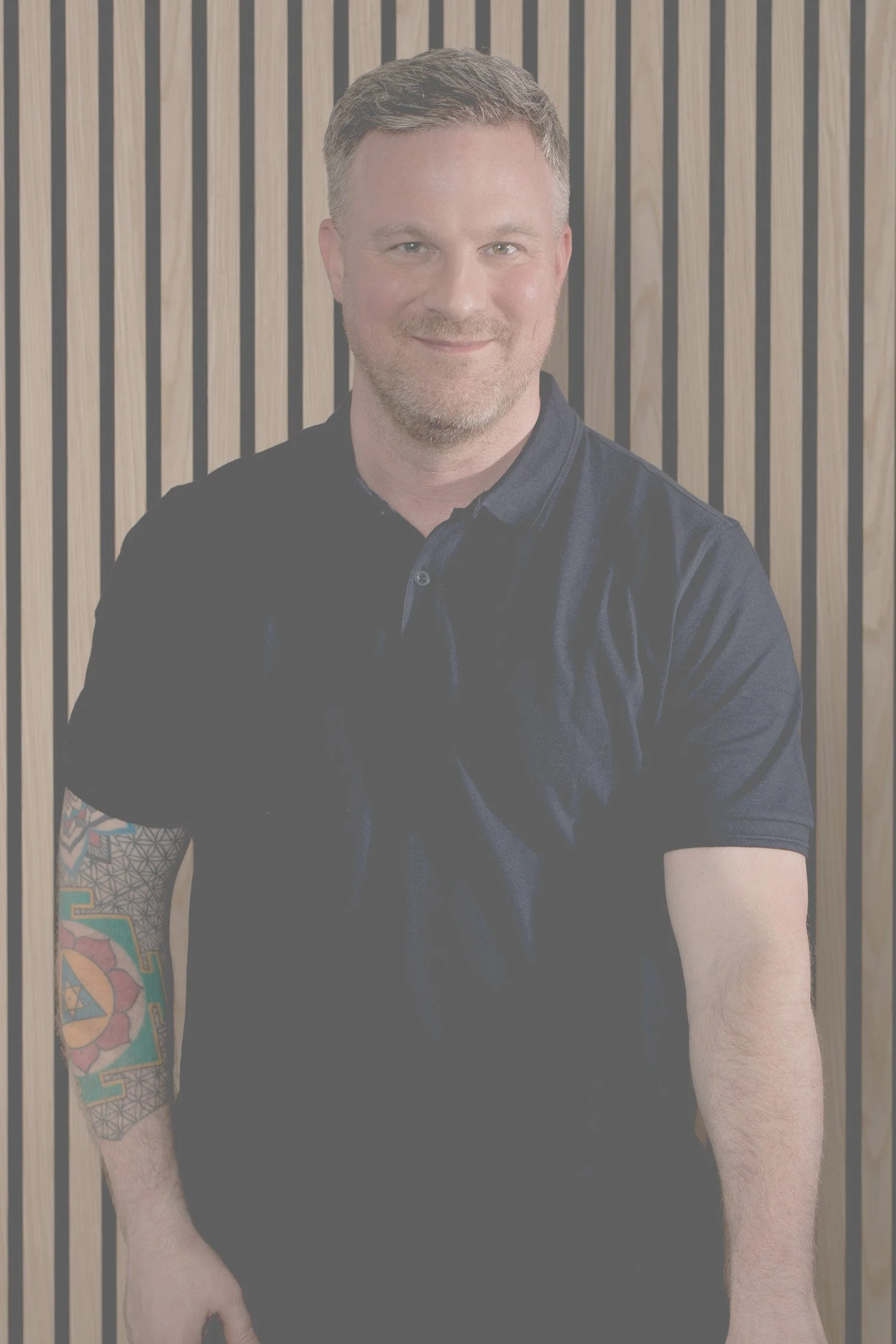 Ein Mann mit kurzem braunem Haar, lächelnd, trägt ein schwarzes Polo-Hemd und hat ein buntes Tattoo auf dem linken Unterarm, steht vor einer Wand mit vertikalen Holzlatten.