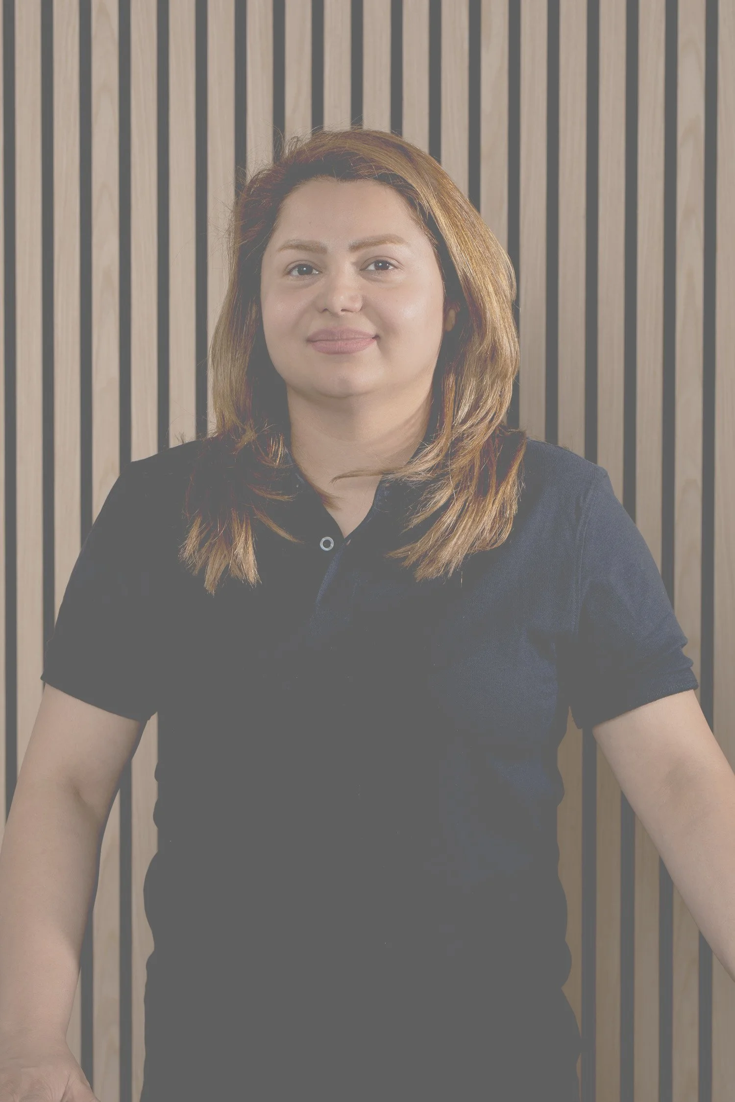 Ein Porträt einer Frau mit braunem, schulterlangem Haar, die einen schwarzen Polohemd trägt, vor einem Hintergrund mit vertikalen Holzlatten.