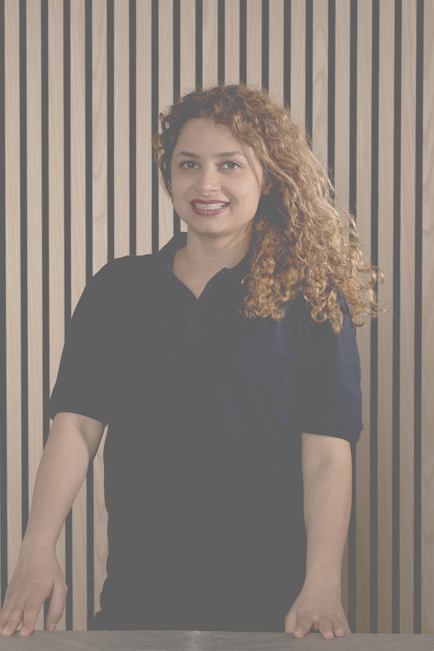 Eine Frau mit lockigem braunem Haar, die an einem Tisch steht, vor einem Holzlamellen-Hintergrund, trägt ein schwarzes Polo-Shirt.