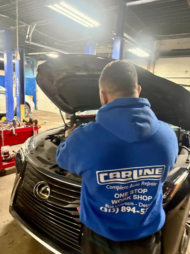 A mechanic in a blue Car Line hoodie working on the engine of a black Lexus inside an auto repair shop with tools and equipment around.