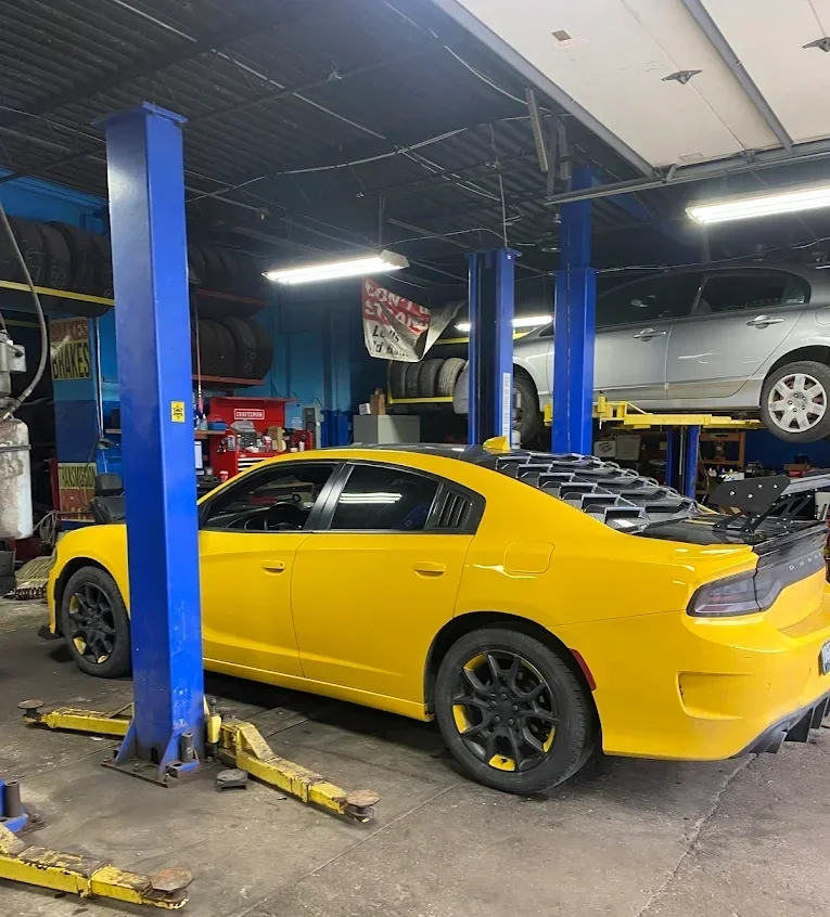 Yellow car inside an auto repair shop with a vehicle lift, and a silver car on an elevated platform in the background.