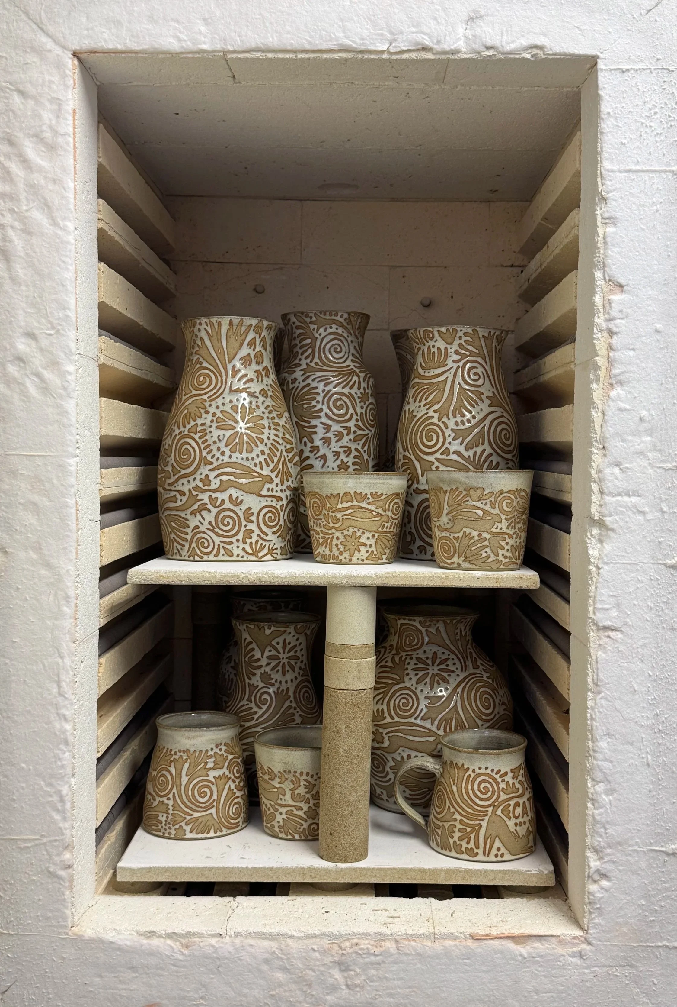 A photo of a finished glaze firing of vases and mugs with a folk-art inspired design of flowers, hares and spirals