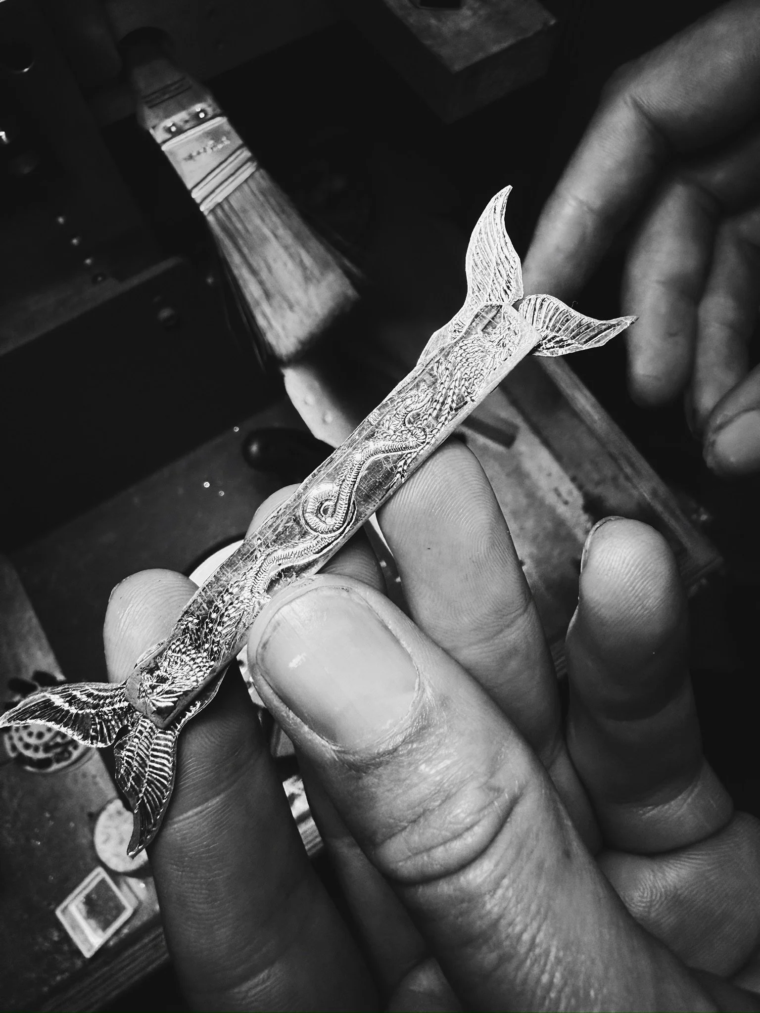 Close-up of a hand holding a detailed, silver fish-shaped object with intricate engravings, with a paintbrush and a paint station in the background.