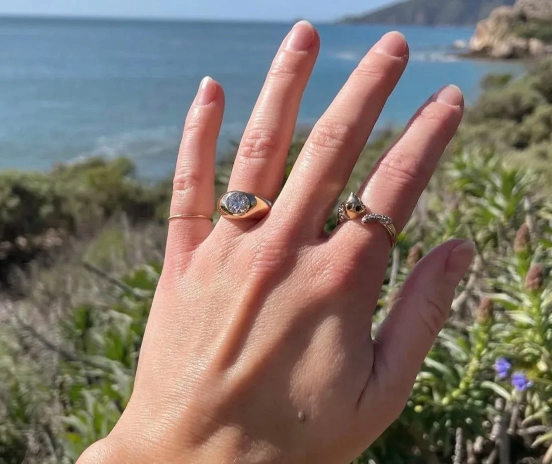 Close-up of a hand with multiple rings, fingers spread, in front of a coastal landscape with water, greenery, and rocky hills.