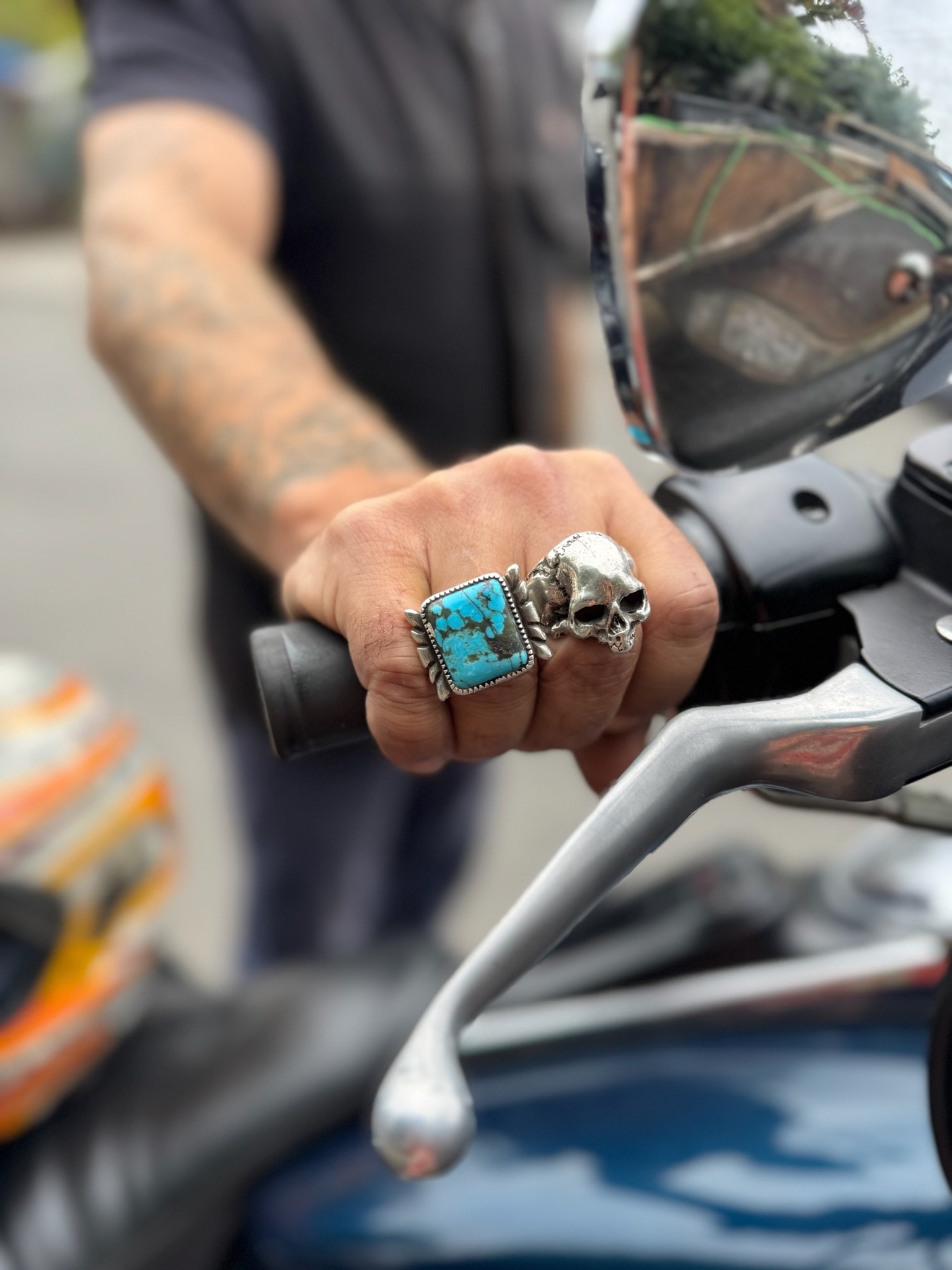 Close-up of a person's hand with a turquoise ring and a skull ring, gripping a motorcycle handlebar.