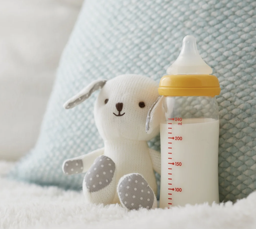 Cute plush toy of a bunny with long ears, sitting next to a baby bottle filled with milk, on a white textured surface with a blue pillow in the background.