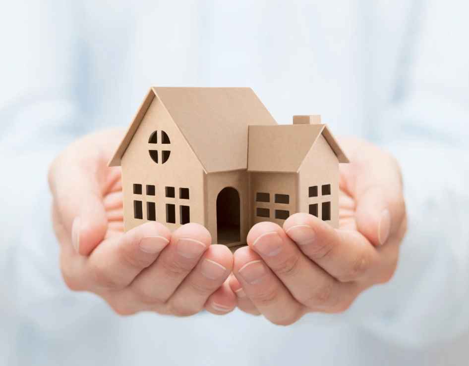 Hands holding a small wooden model house against a blurred background.