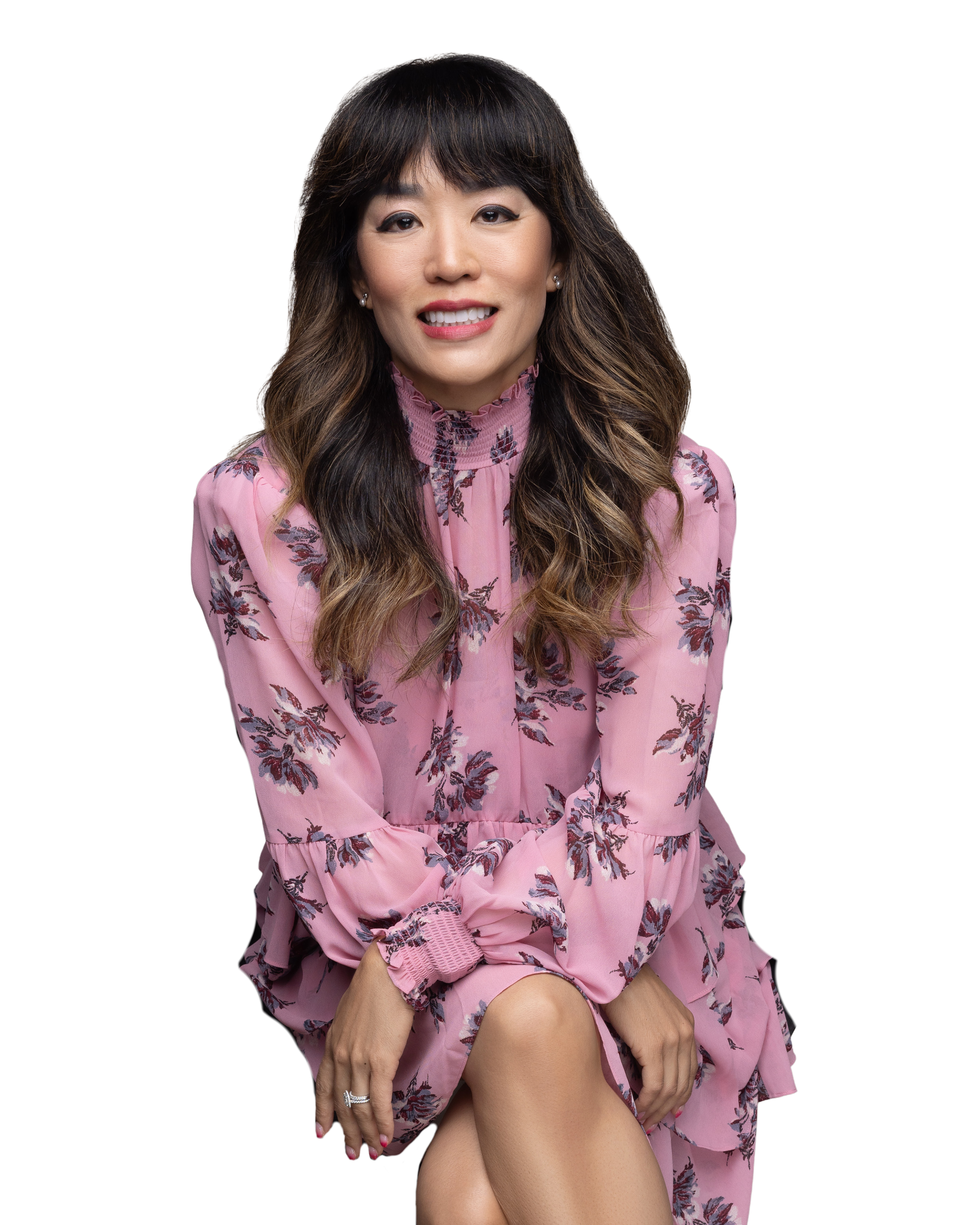 A woman with long wavy hair, wearing a pink floral dress, sitting with her legs crossed, smiling at the camera.