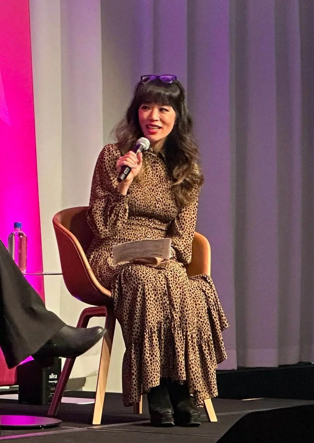 Coach Andrea Chang, a woman with long dark hair wearing a light brown dress and black boots, is sitting on a brown chair, holding a microphone, and speaking on stage with gray curtains in the background.