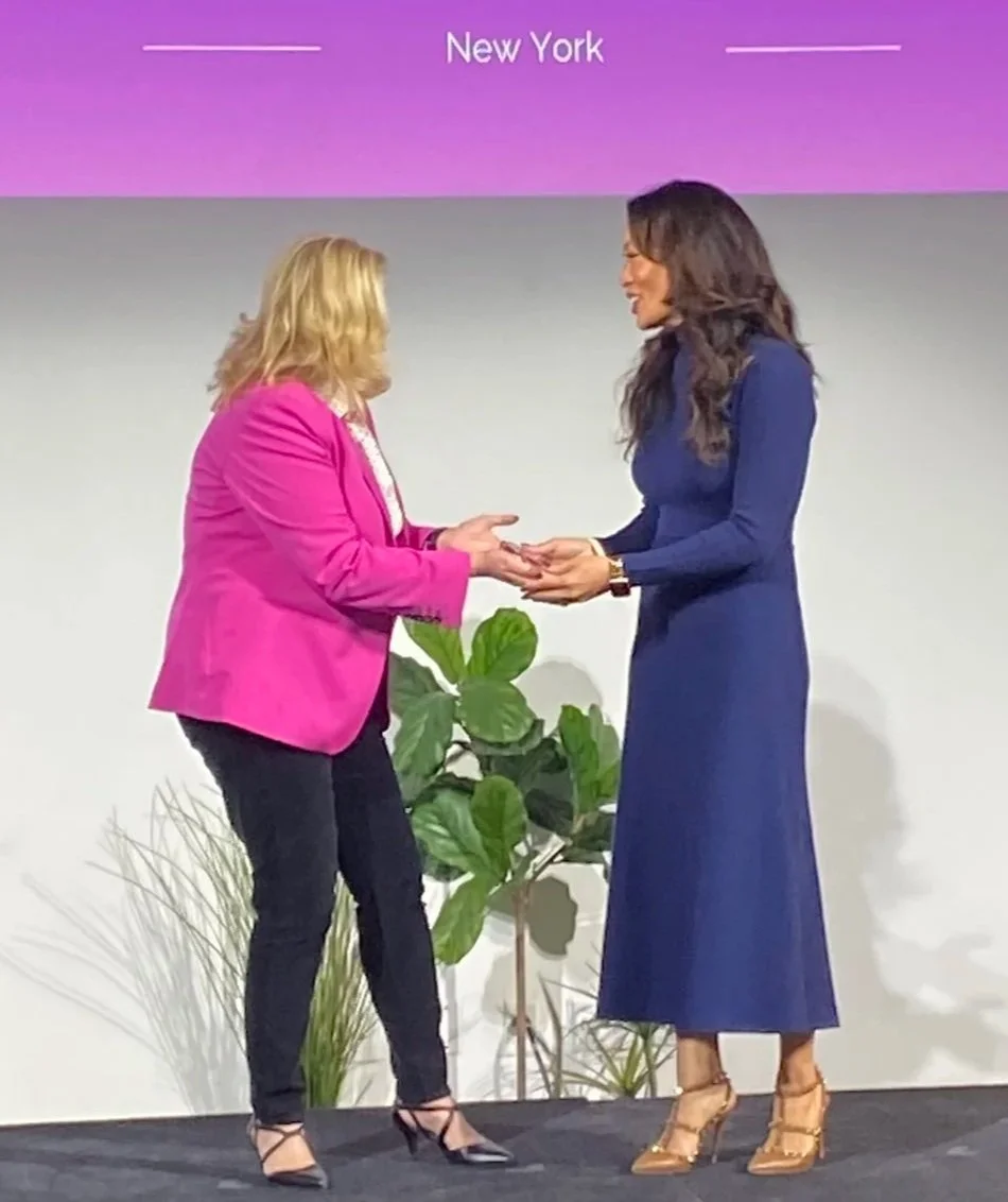 Two women exchanging an award on stage, with a plant in the background and 'New York' written on the screen above.