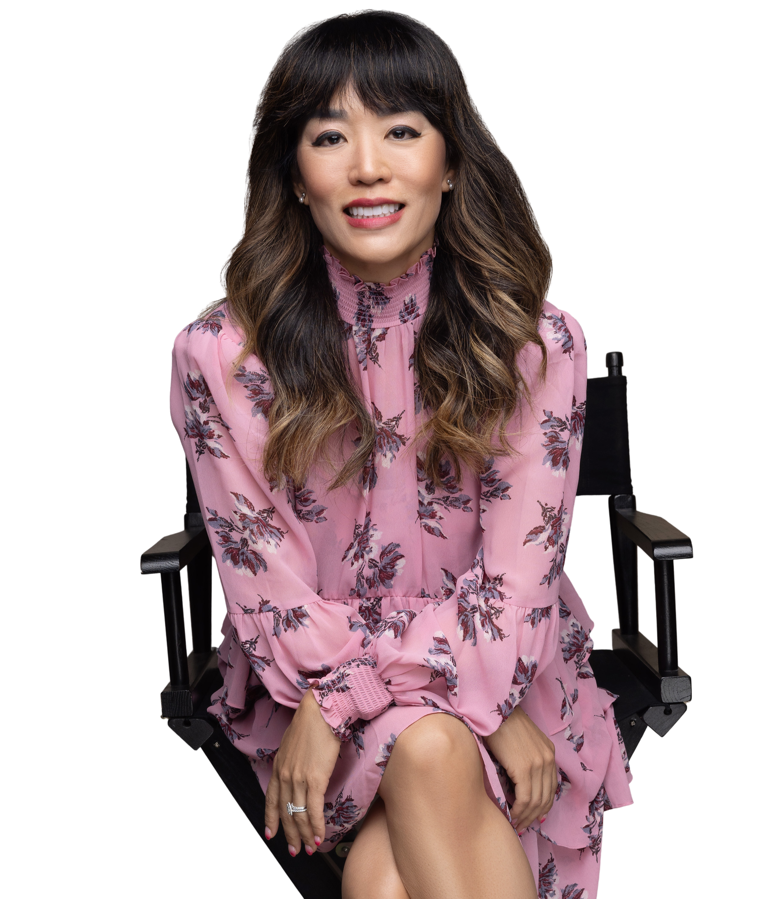 Coach Andrea Chang, a woman with long dark hair, is sitting on a black director's chair, wearing a pink floral dress, smiling at the camera against a transparent background.