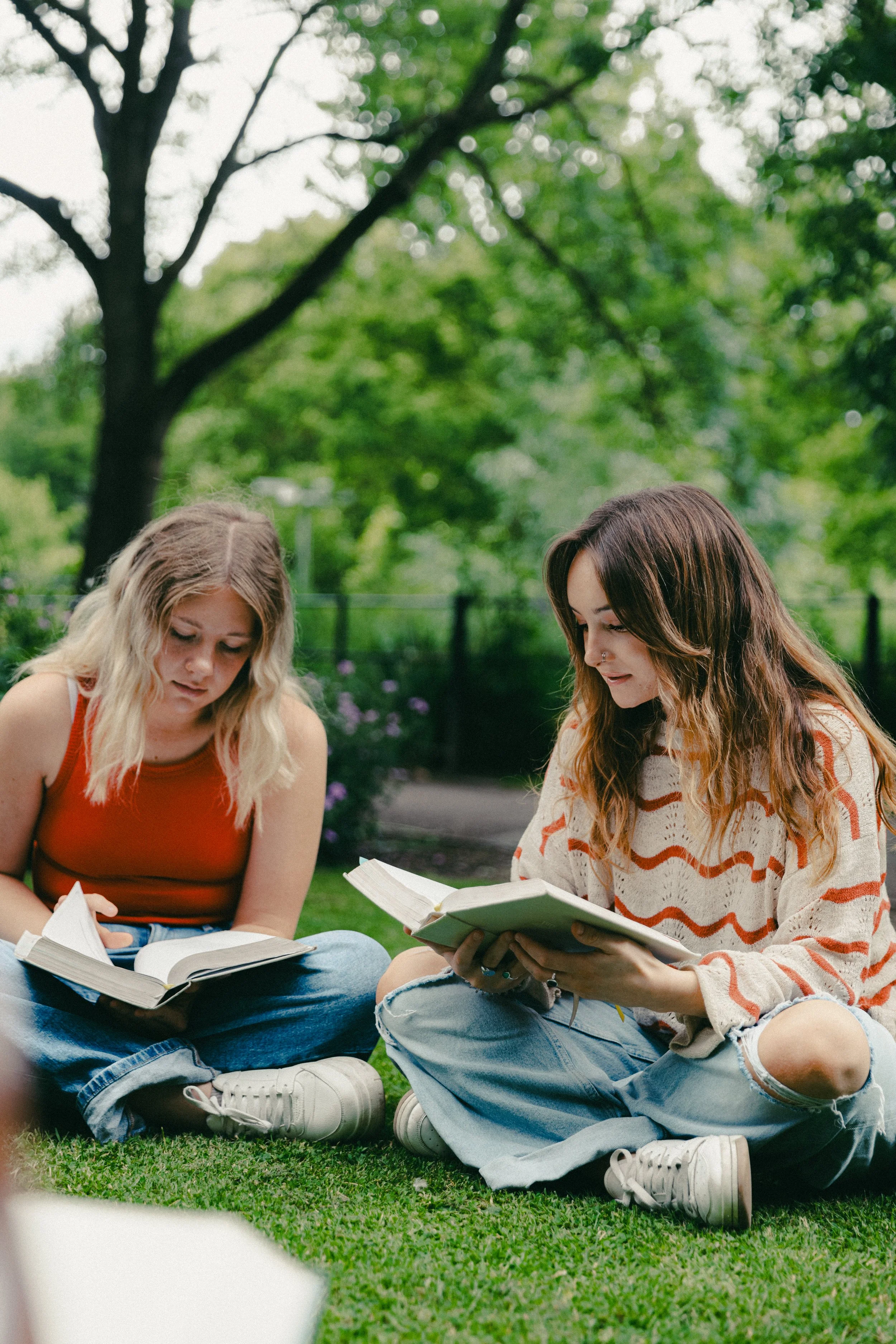 Two young women sitting on the grass in a park, reading books, surrounded by green trees and plants on a cloudy day.