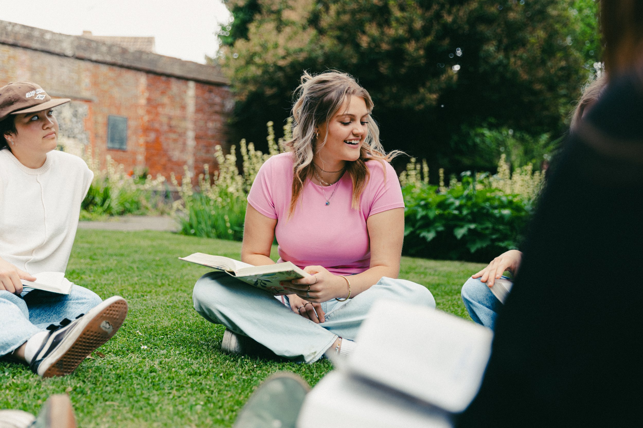 Young woman in pink shirt sitting on grass, reading a book, smiling, with other people nearby in an outdoor park.