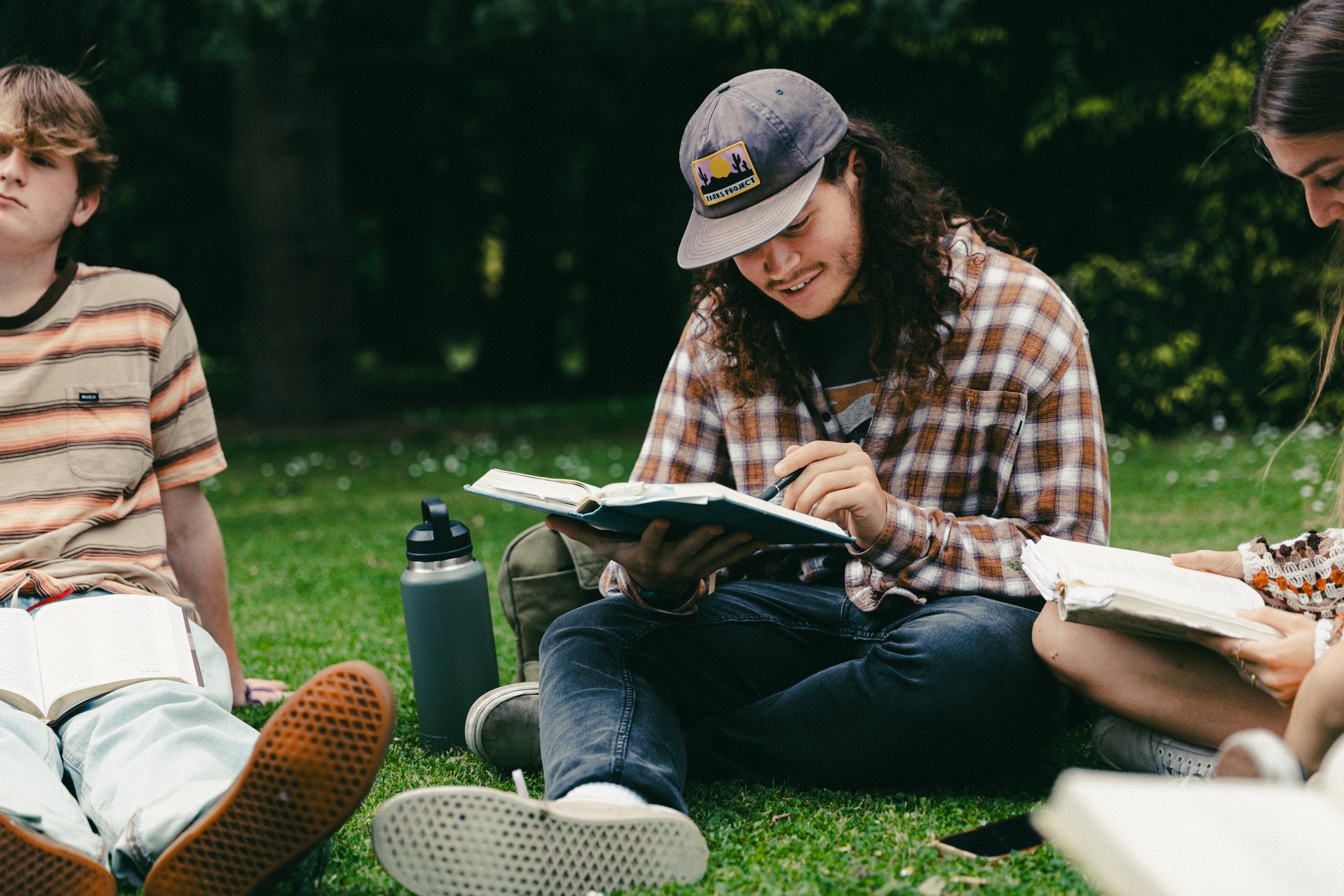 Group of young people sitting on grass outdoors, engaged in studying or reading with notebooks and books around them.