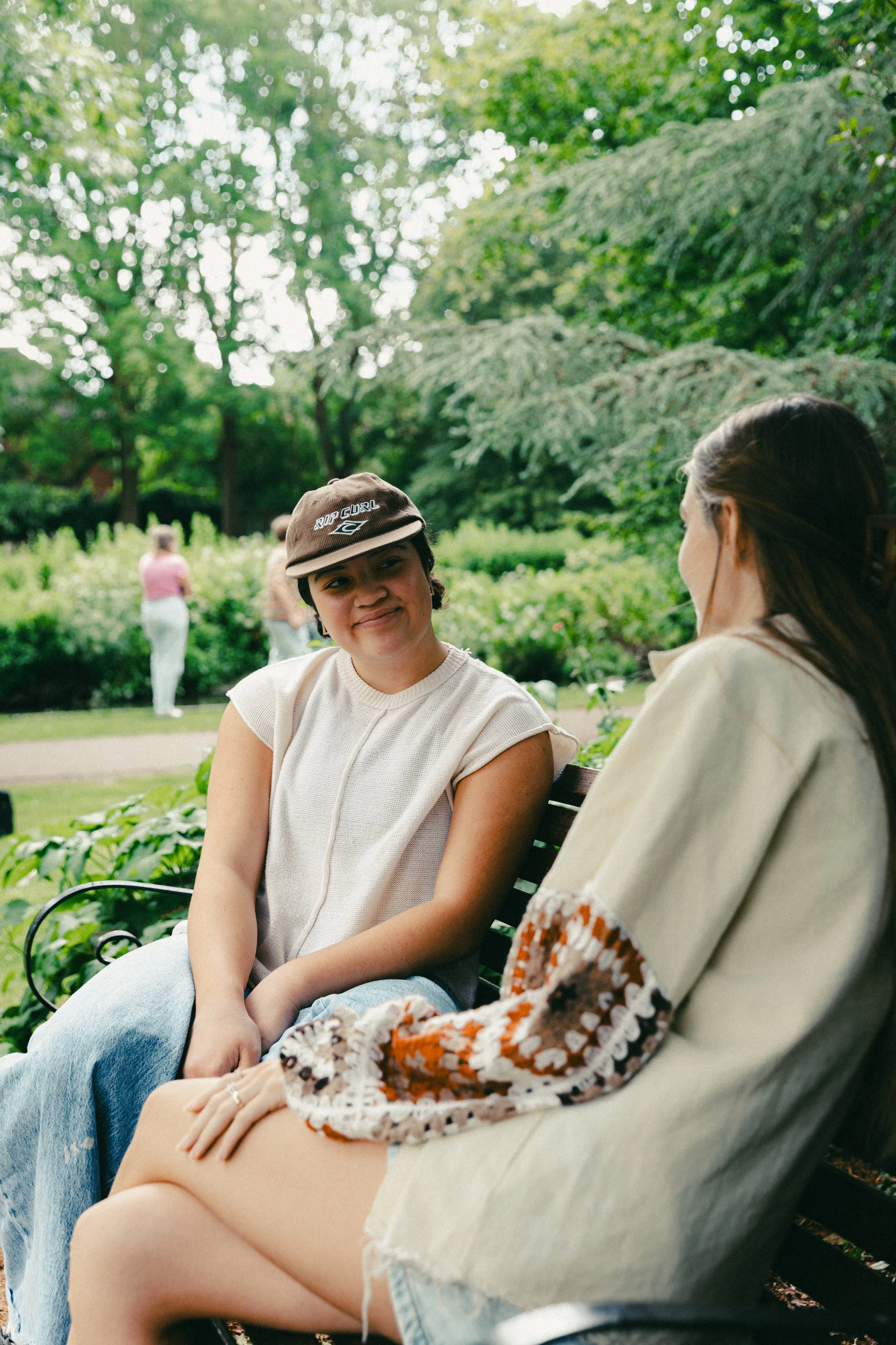 Two women sit and converse on a park bench, with lush green trees and plants in the background.