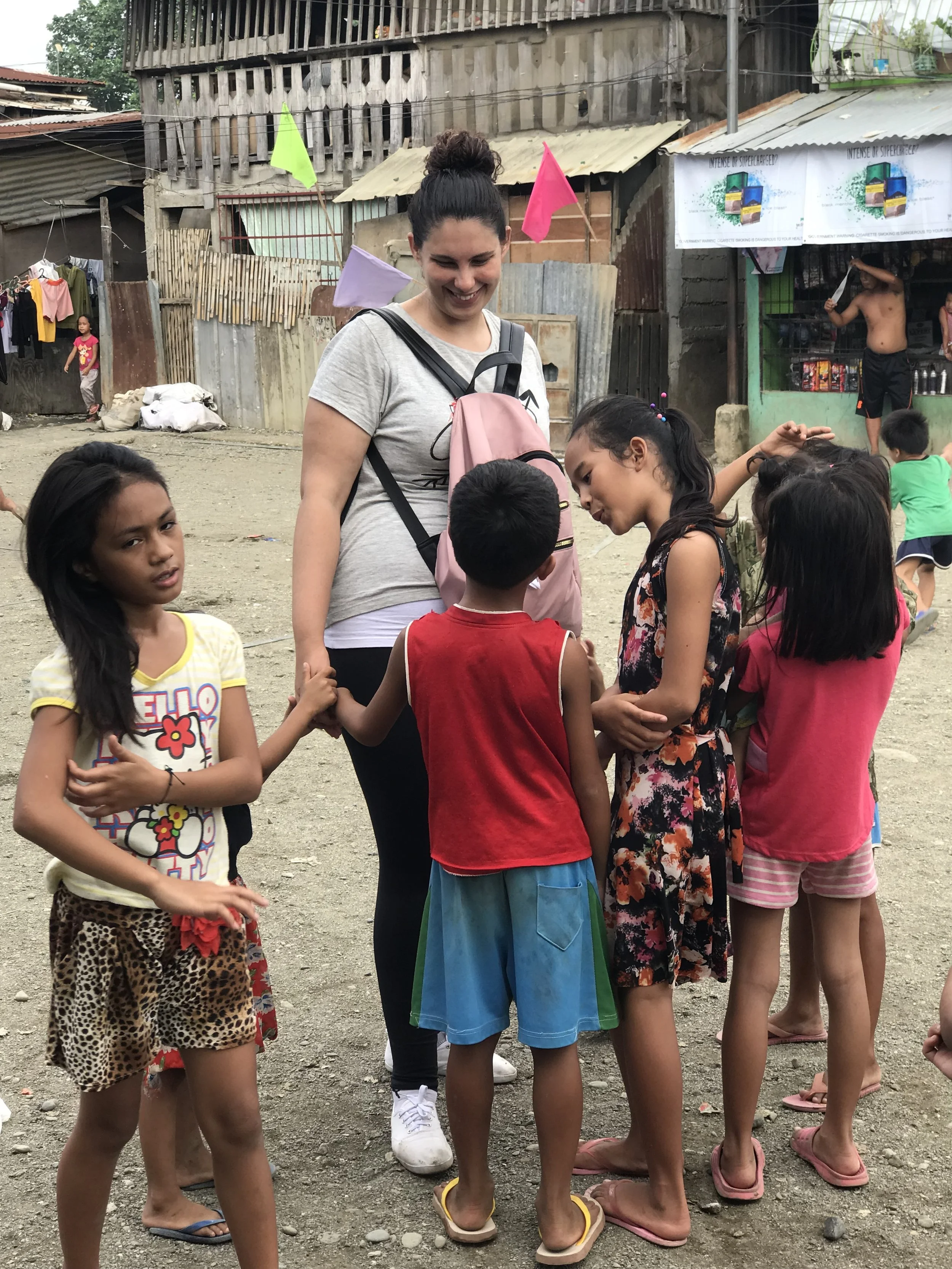 A woman with a pink backpack smiling and holding hands with a group of children in a dusty outdoor area with makeshift buildings and other children playing in the background.
