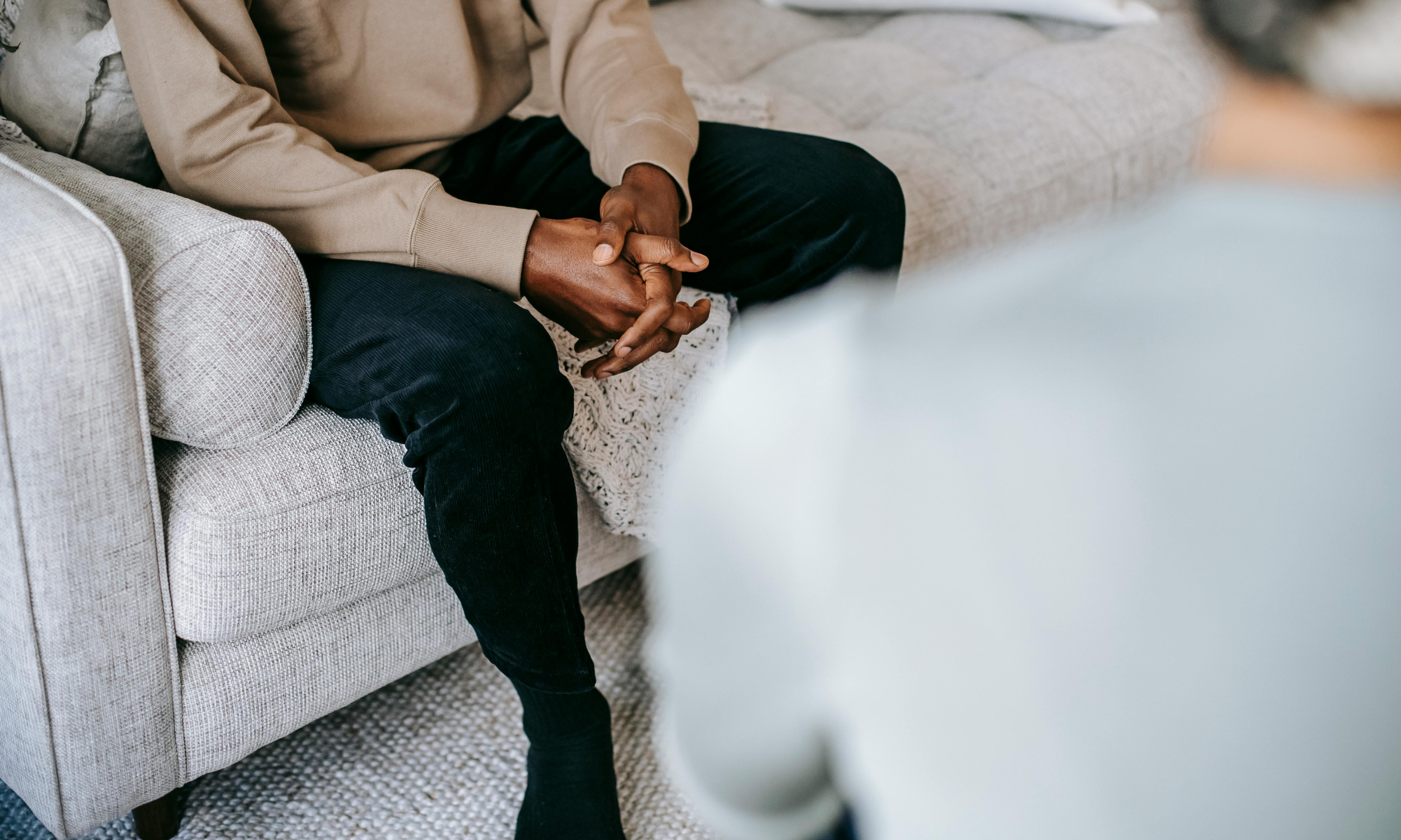 A person sitting on a beige fabric sofa with their hands clasped together on their lap, wearing a tan sweatshirt and black pants, during a conversation with another person whose shoulder is partially visible in the foreground.