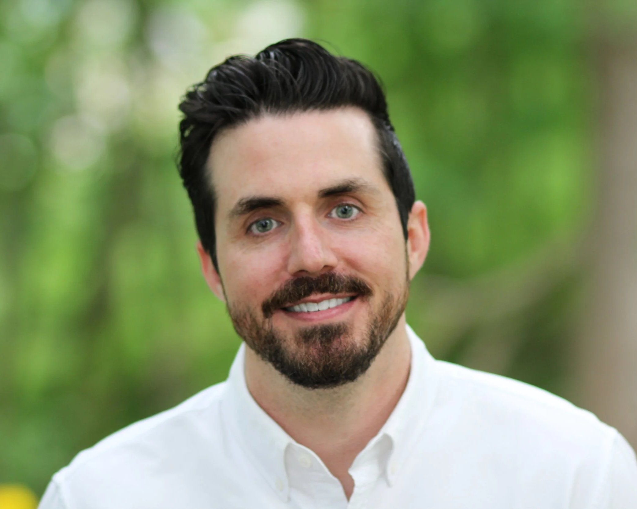 A man with black hair, a beard, and mustache, wearing a white shirt, smiling outdoors with a blurred green background.