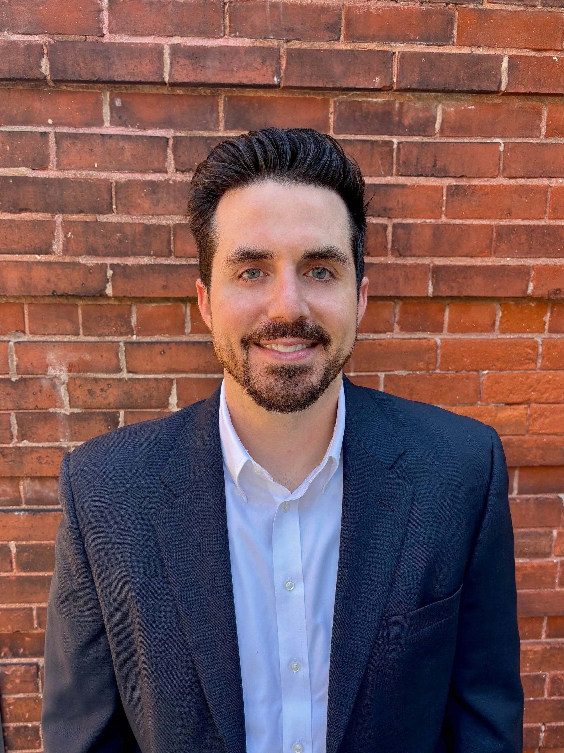A man with dark hair, a beard, and blue eyes wearing a navy suit and white shirt, standing in front of a brick wall.