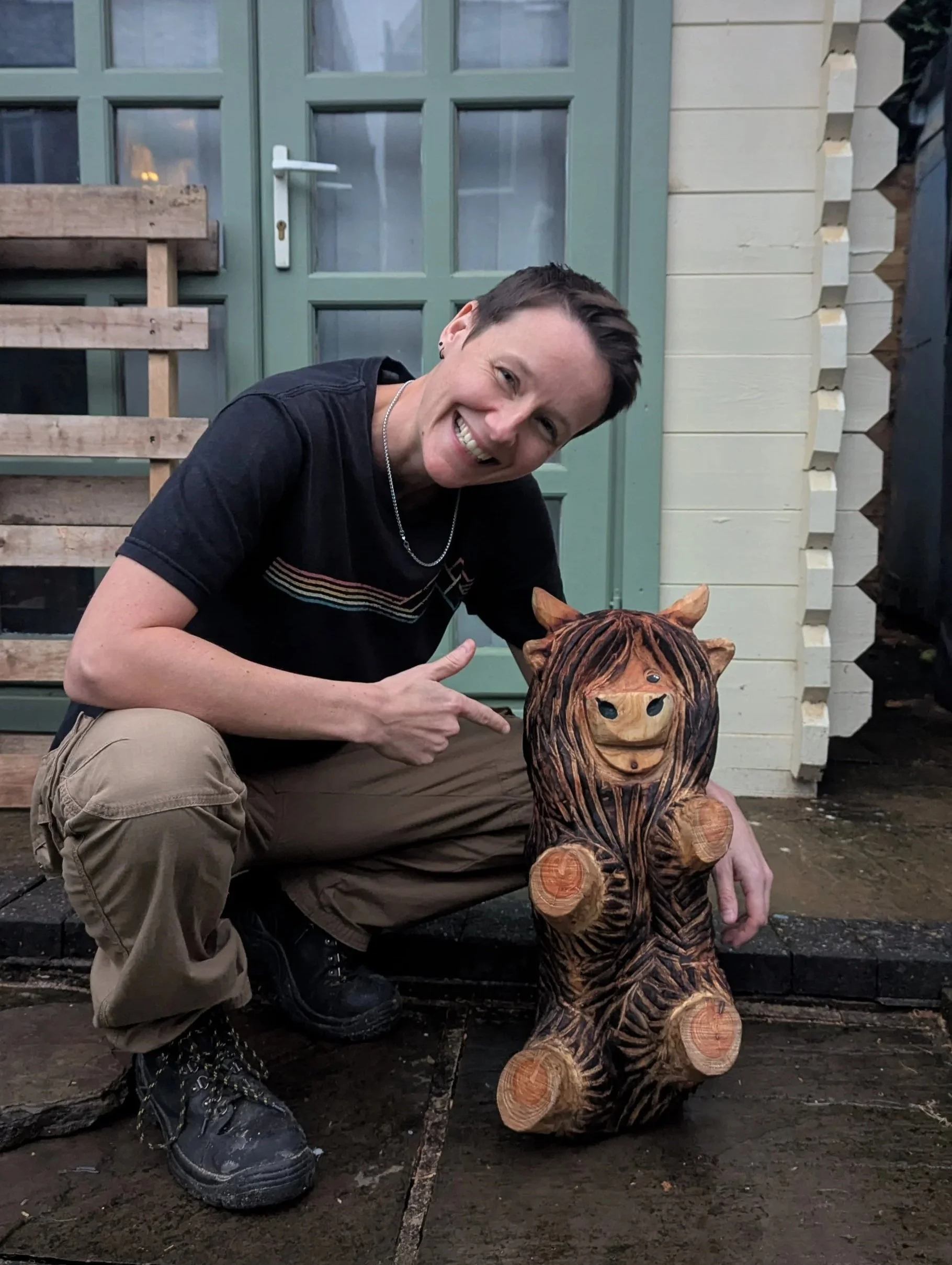 A smiling person crouching next to a wooden highland cow sculpture outside a house with green door.