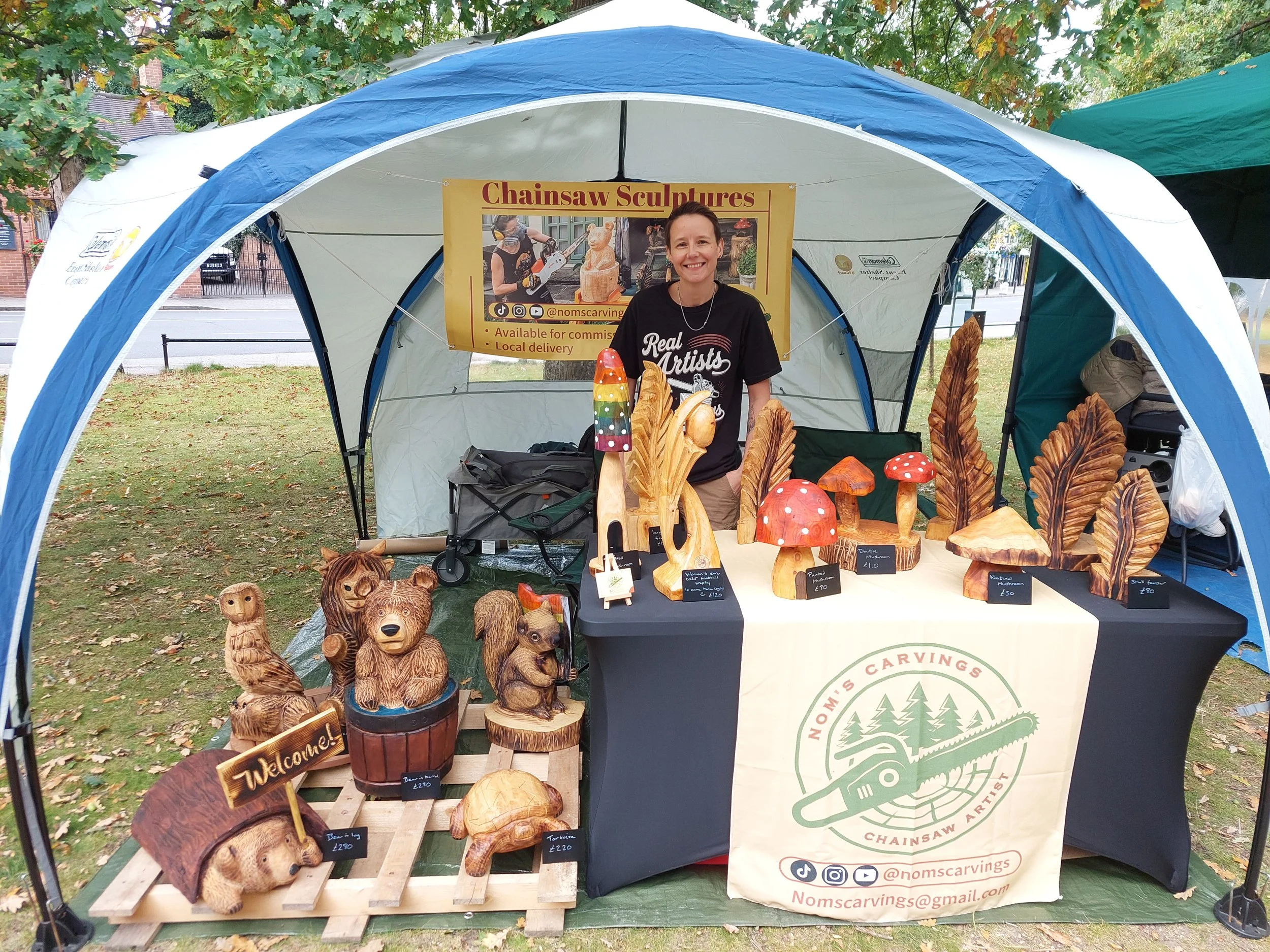 Woman standing behind a table displaying wooden carvings at an outdoor craft fair. The carvings include bears, mushrooms, leaves, an owl, a fox, and a turtle. A sign above advertises "Chainsaw Sculptures" and mentions local delivery. The table has a beige cloth with a logo and contact information for Noms Carvings.
