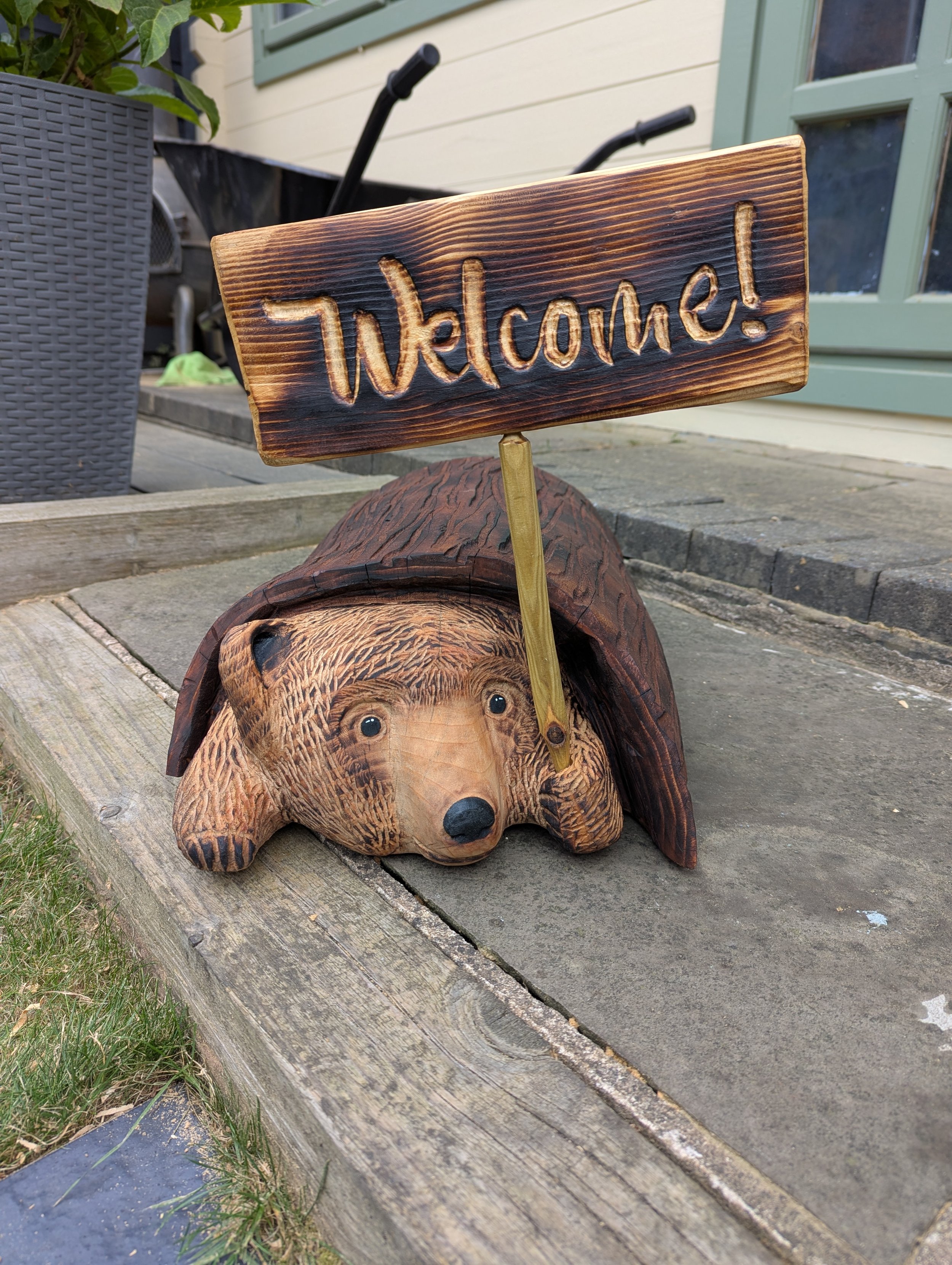 A decorative wooden bear sculpture lying on the ground with a small wooden sign on top that says "Welcome!"
