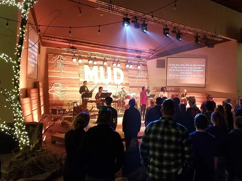 A church service with a band performing on stage with the word 'HUMD' in large lights behind them. The congregation is standing and singing, with lyrics projected on a screen. The decor includes string lights and wooden panels.