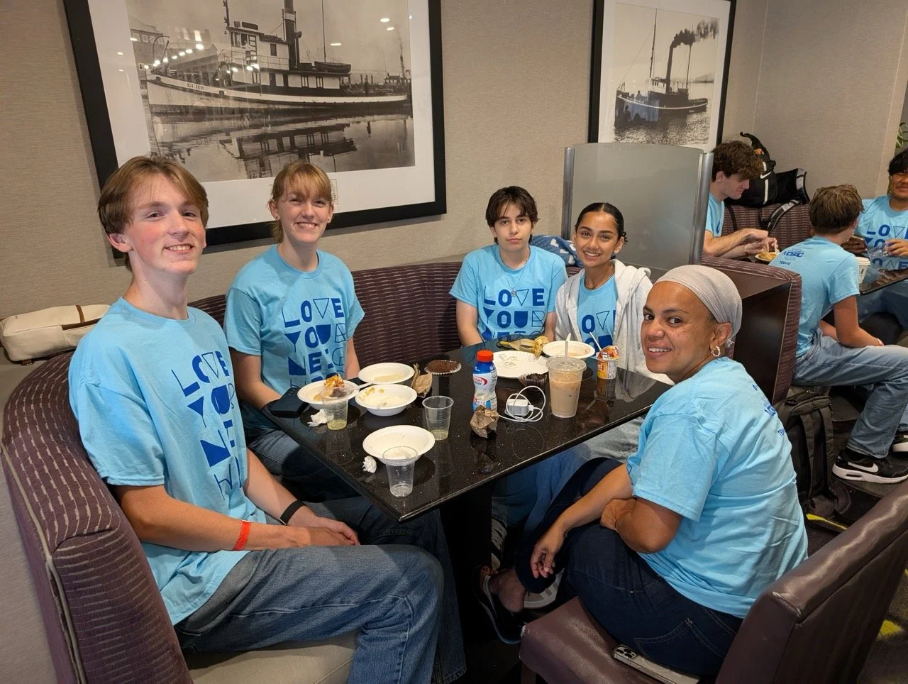 Group of six young people sitting around a table in a restaurant or cafe, smiling at the camera. They are wearing matching light blue T-shirts with blue text, and there is food and drinks on the table. Framed black and white photos of ships hang on t