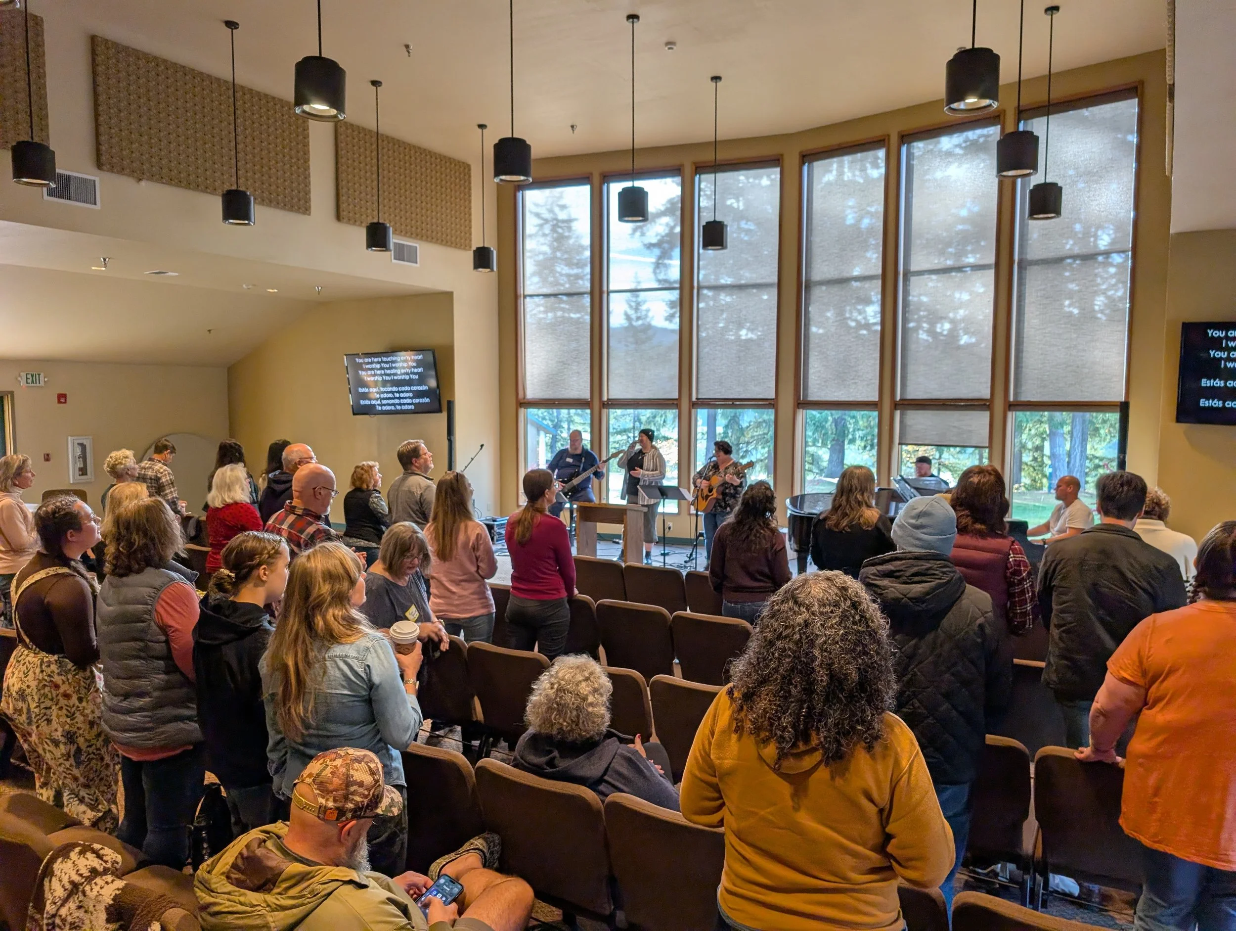 People gathered in a chapel or auditorium listening to a live musical performance with three band members playing drums, guitar, and keyboard near large windows.
