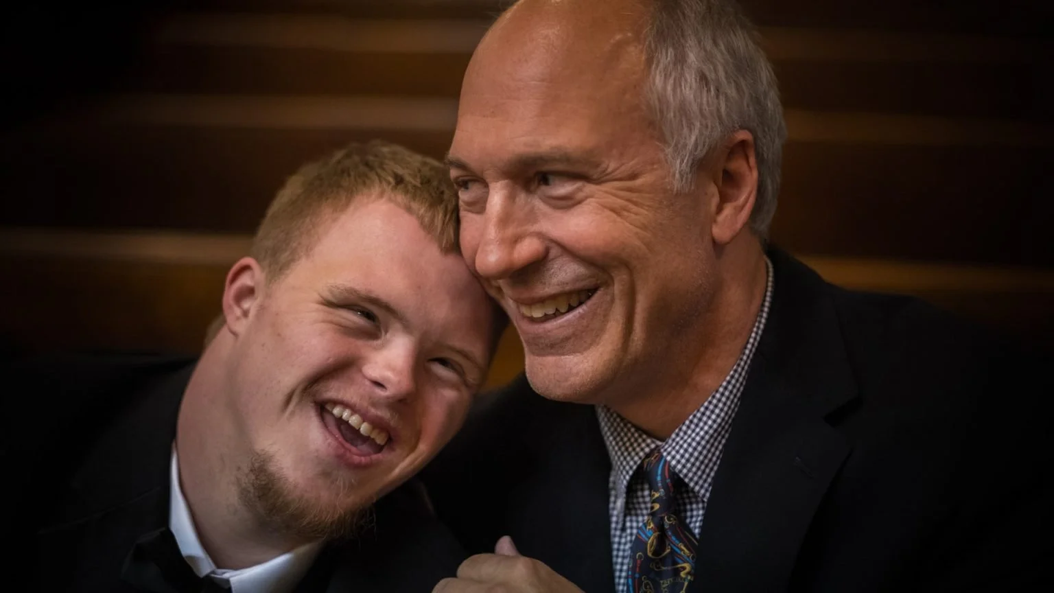 Two men sharing a joyful moment, one leaning his head on the other's shoulder, both smiling and laughing.