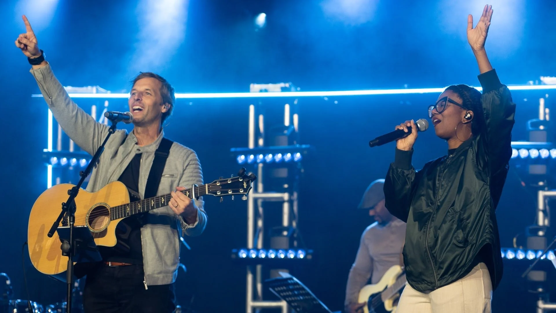 Two musicians on stage, a man playing guitar and singing into a microphone, and a woman singing into a microphone with her hand raised, surrounded by bright stage lights.