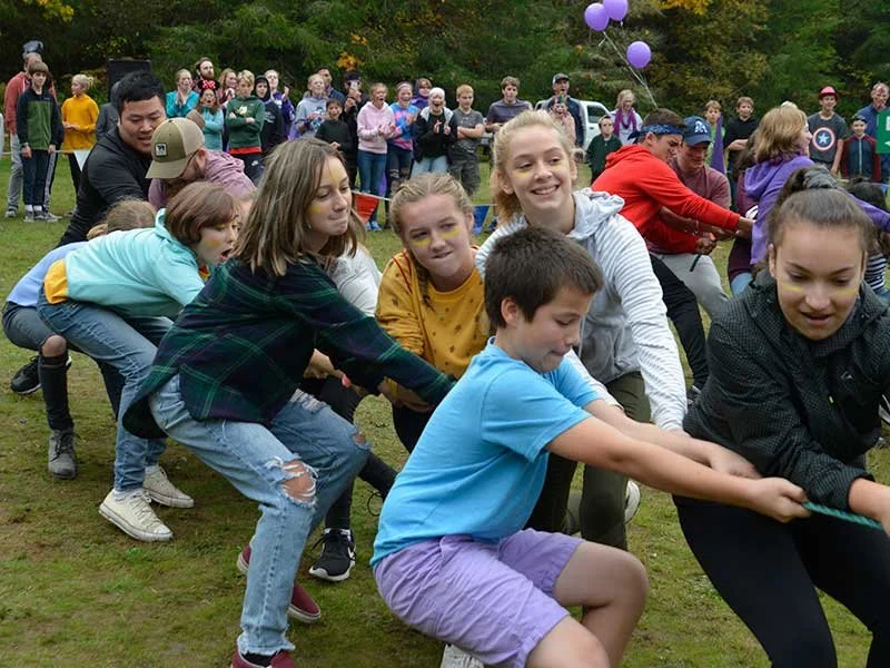 Children and adults participating in a tug-of-war game outdoors in a grassy area, with trees and balloons in the background.