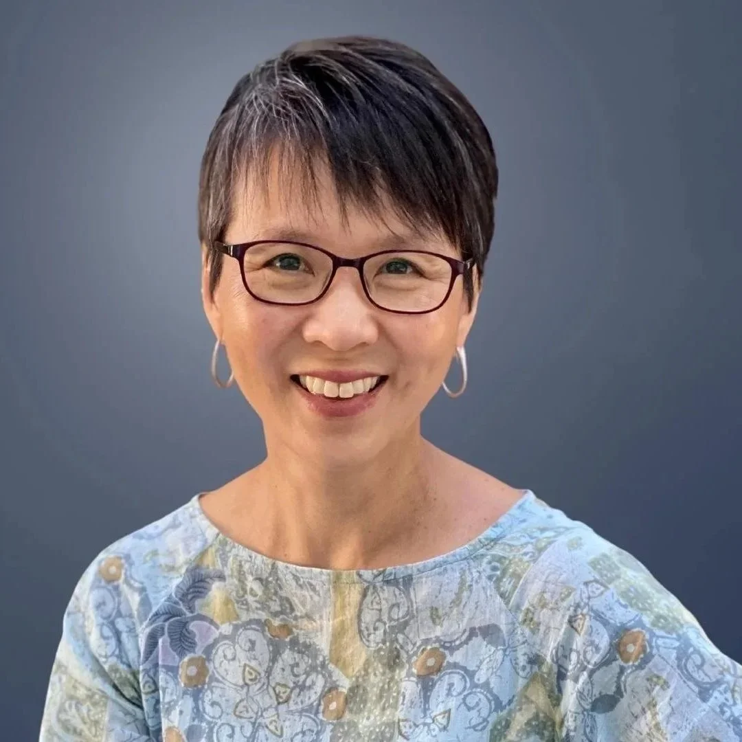 A smiling woman with short dark hair, glasses, and hoop earrings, wearing a patterned light-colored top, against a plain gray background.