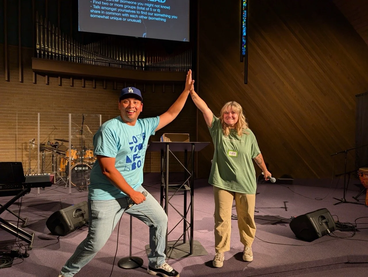 Two people celebrating with a high five on stage in a church or event hall, holding microphones, with music instruments in the background, and a large screen displaying text above them.