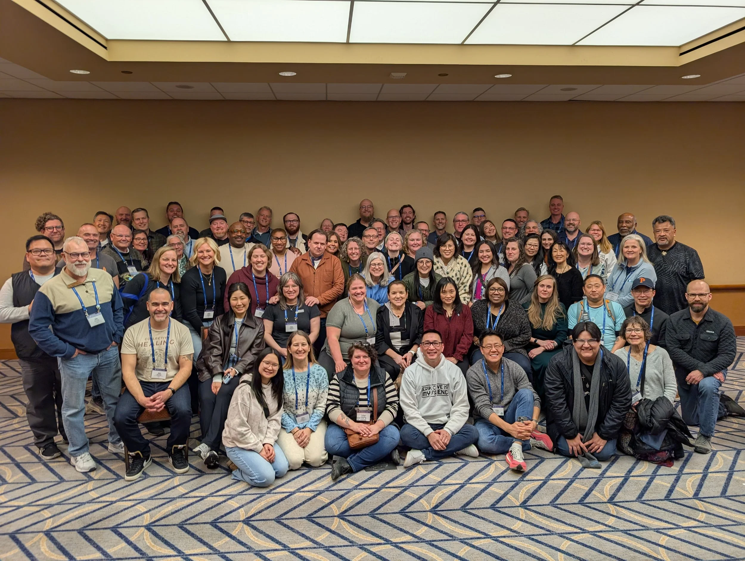 Large group of diverse people posing for a photo in a conference room with a beige wall and patterned carpet.