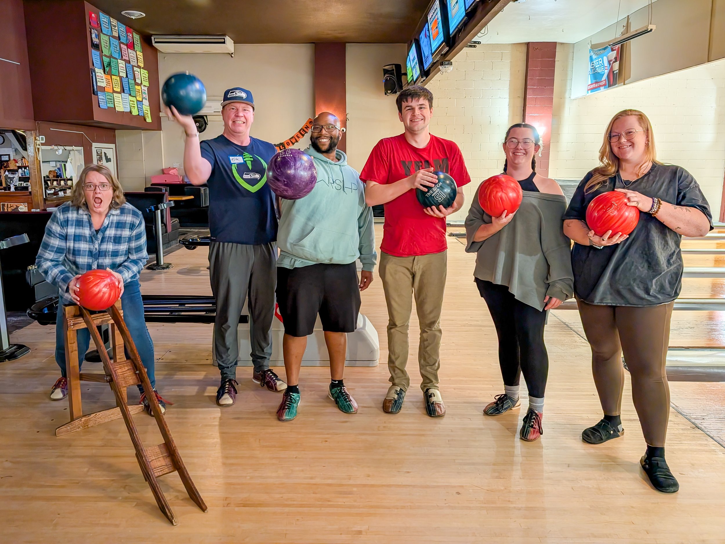 Seven people at a bowling alley holding bowling balls, smiling for the camera. One person on the far left is standing with a red ball, second person from the left is holding a blue ball, third person with glasses holds a purple ball, fourth person in a red shirt holds a black ball, fifth person in a gray sweatshirt holds a red ball, sixth person in a black shirt also holds a red ball. There is a wooden chair with a red bowling ball on it and a bowling lane in the background.