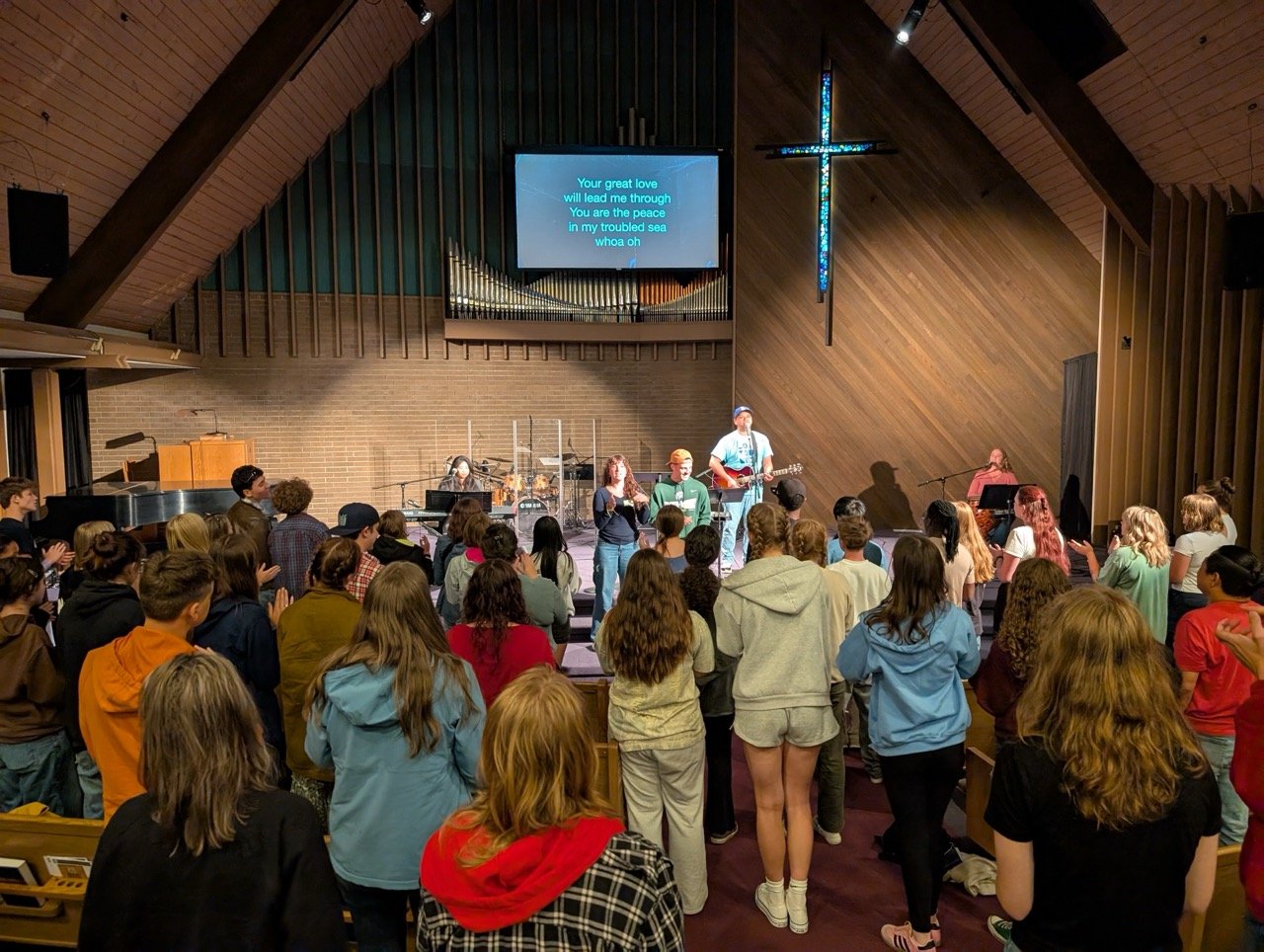 People gathered in a church interior for a service or event, with a band performing on stage, a large cross on the wall, and lyrics on a screen.