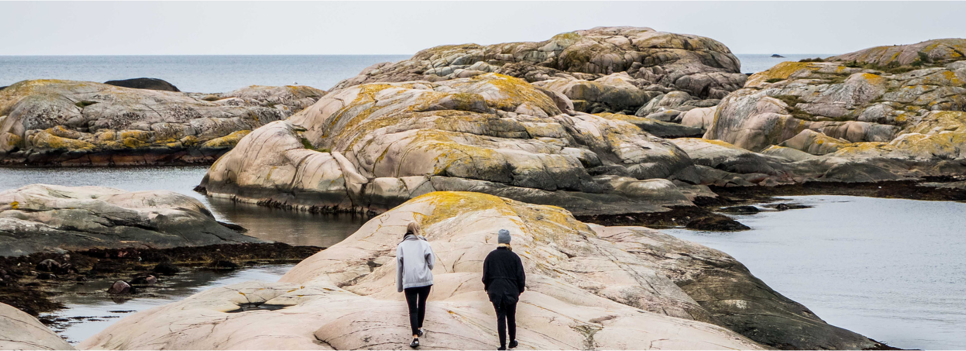 Two people walk on a rocky shoreline near the ocean, with large, weathered rocks extending into the water and overcast sky.