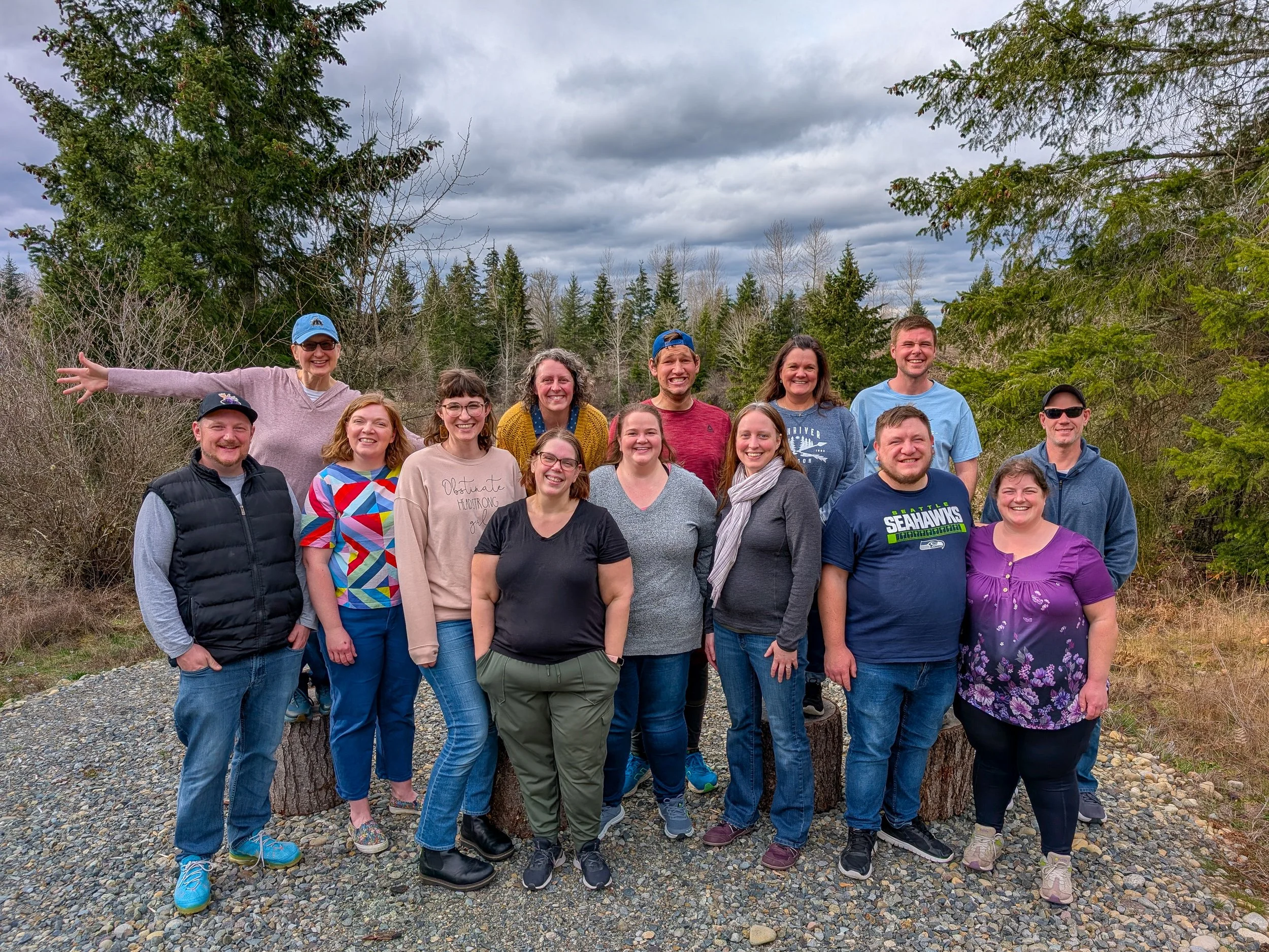 Group of fifteen people outdoors on a cloudy day, standing among trees and rocks, smiling and posing for a photo.