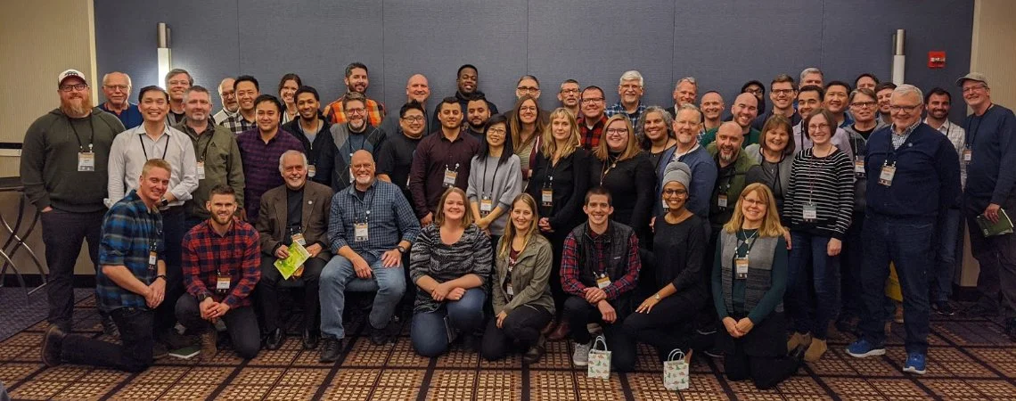 Group of diverse people posing for a photo in a conference room, some holding conference badges and tote bags, smiling at the camera.