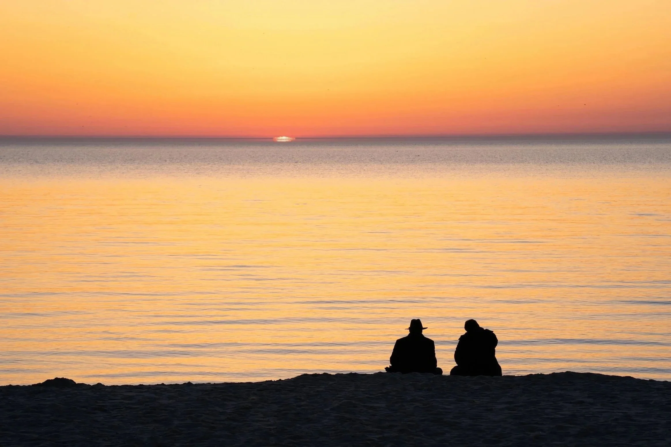 Two people sitting on a beach at sunset, looking at the water and the sunset over the horizon.