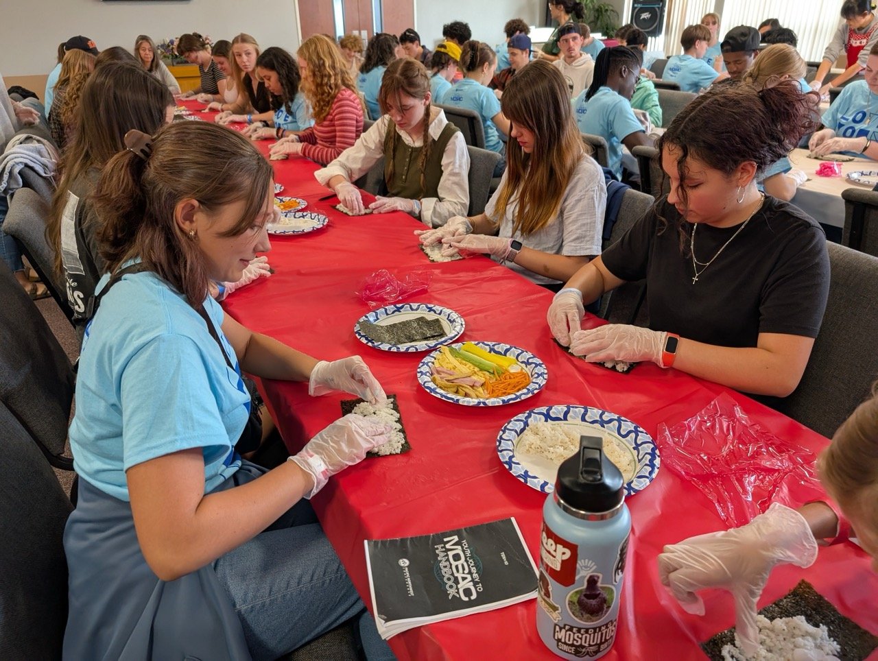 Group of people sitting at a long table covered with a red tablecloth, wearing gloves, preparing sushi rolls with various ingredients on plates in front of them.