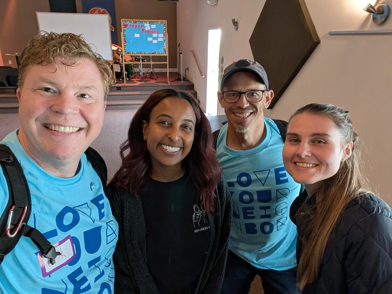 Four smiling people taking a selfie inside a church or community center. There are stage stairs and a colorful bulletin board with words like 'Prayer' and 'Praises' in the background.